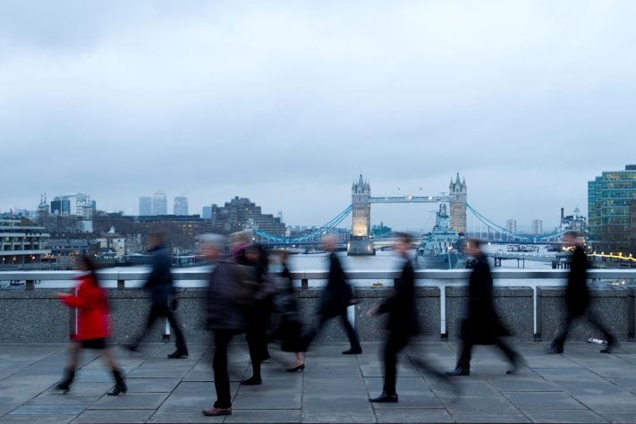 Pedestrians in business attire walk across a riverside walkway, with London’s Tower Bridge visible in the background under an overcast sky.